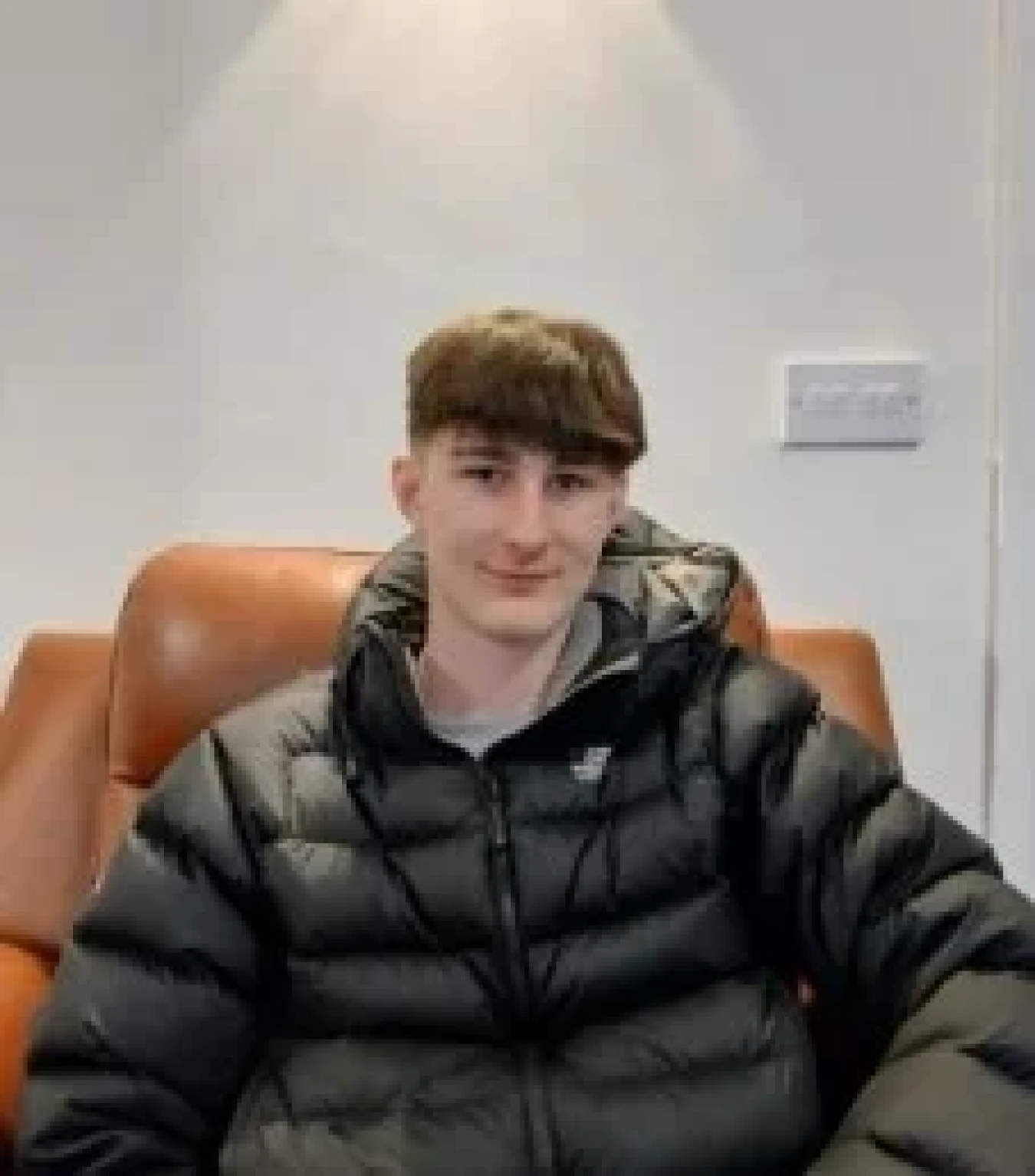 Young man sitting on a brown leather couch wearing a black puffer jacket indoors