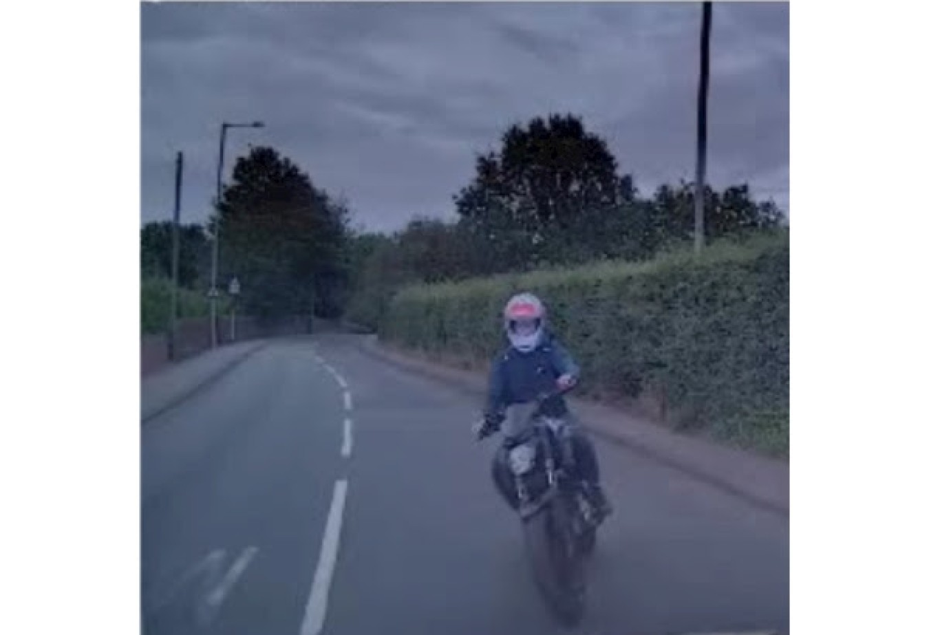 Motorcyclist riding on a suburban road with hedges, wearing a helmet and protective gear.