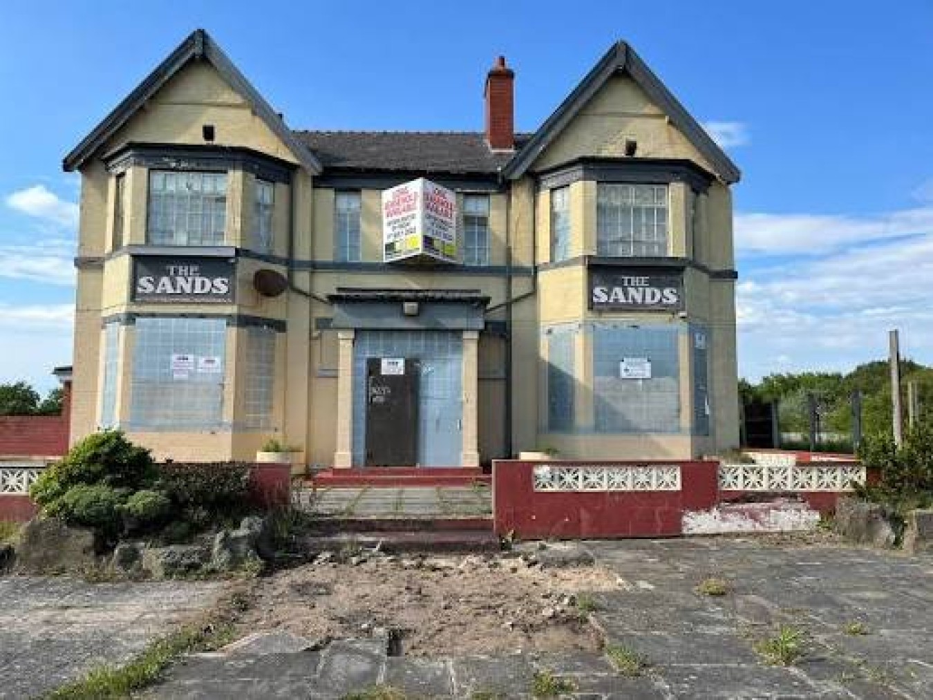 A two-storey building with bay windows, boarded-up doors and signs reading 'The Sands' on both sides, set in a derelict state.