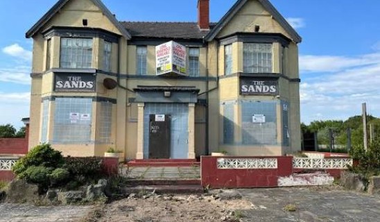 A two-storey building with bay windows, boarded-up doors and signs reading 'The Sands' on both sides, set in a derelict state.