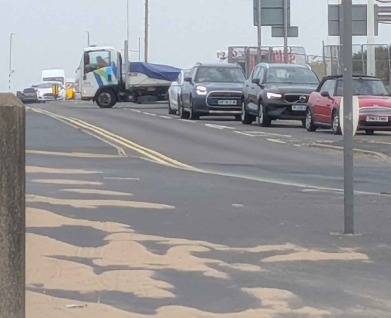 Traffic queue on a coastal road with a white van and several cars awaiting ahead, road markings and a lamp post in view.
