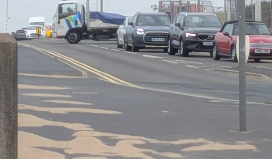 Traffic queue on a coastal road with a white van and several cars awaiting ahead, road markings and a lamp post in view.