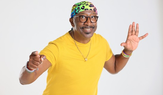 Smiling man wearing a bright yellow shirt and multicoloured headscarf, reaching towards the camera.