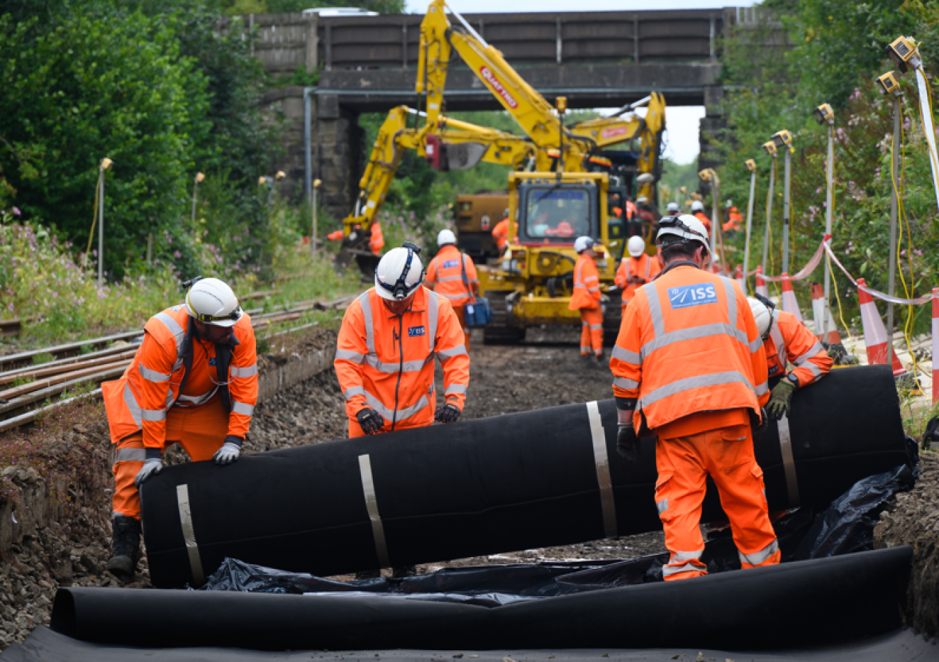 construction workers in orange overalls lay down large black piping in a trench near a railway underpass
