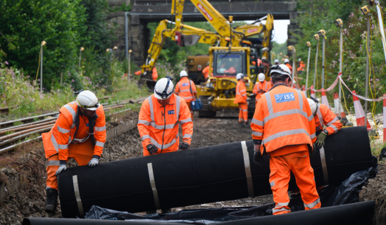 construction workers in orange overalls lay down large black piping in a trench near a railway underpass