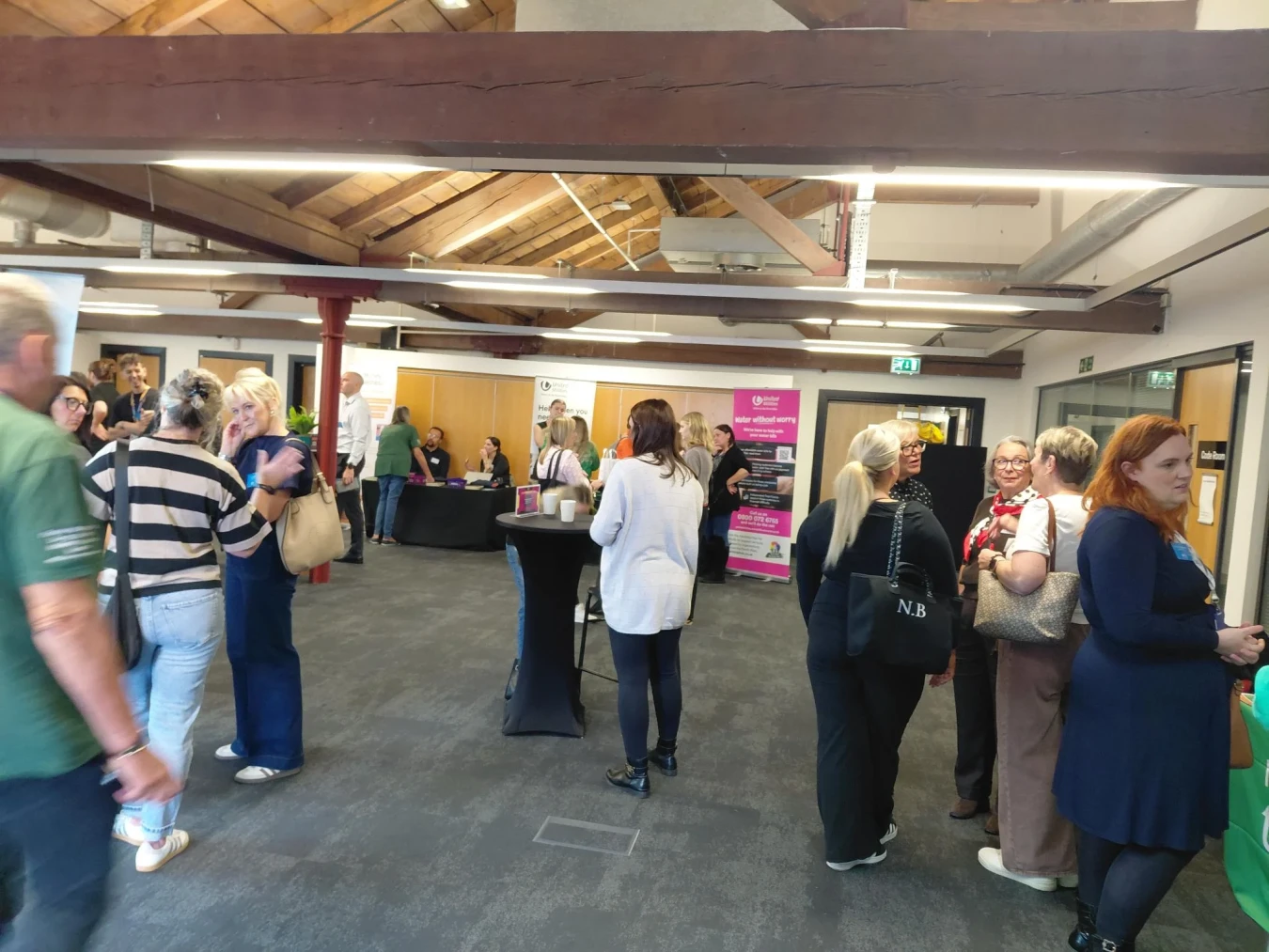 Busy indoor event with people chatting near stalls and banners under wooden beams.