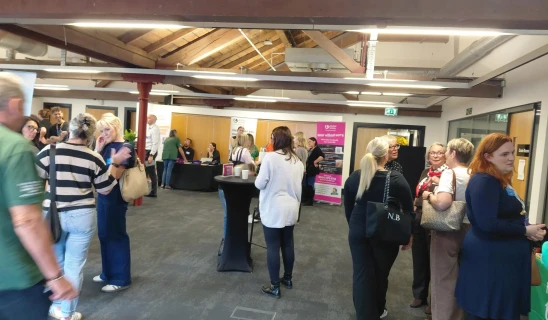 Busy indoor event with people chatting near stalls and banners under wooden beams.