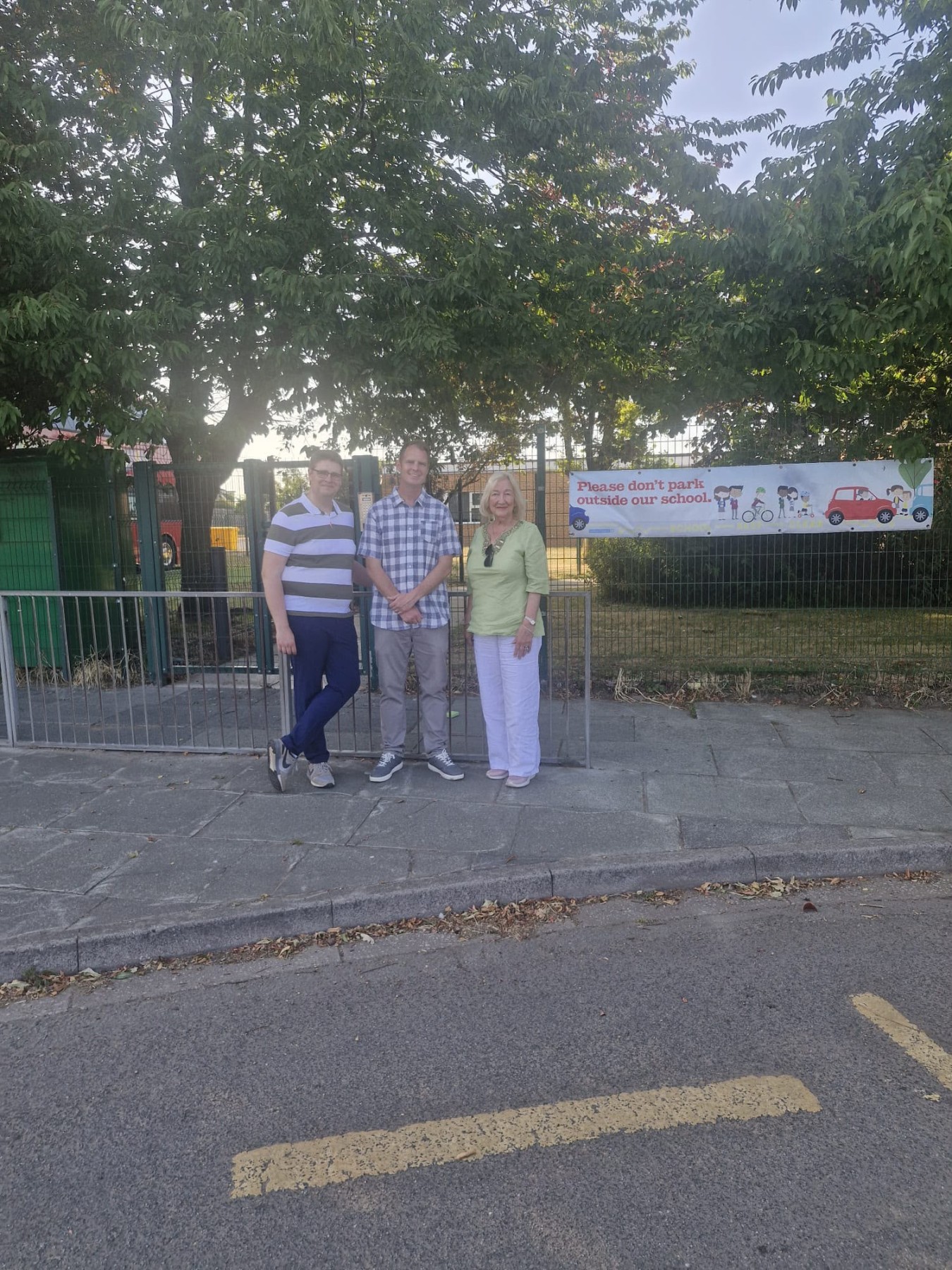 Three adults stand by a metal fence with a banner in the background reading, Please don't park outside our school.