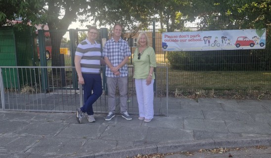 Three adults stand by a metal fence with a banner in the background reading, Please don't park outside our school.