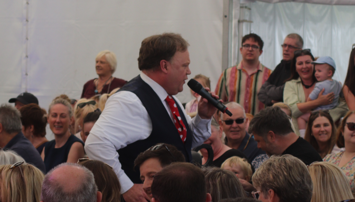 Man in white shirt and red polka dot tie singing into microphone at crowded event.