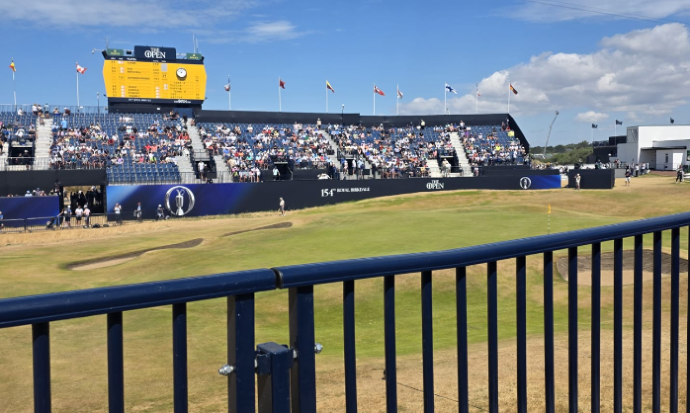 Cricket stadium with blue seating, scoreboard and spectators under blue sky, foreground railing