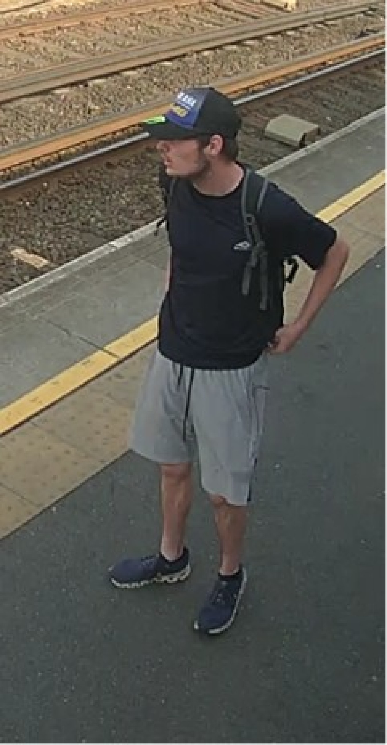 young man standing on train platform, wearing cap, black t-shirt, light shorts and sneakers, looking to the side