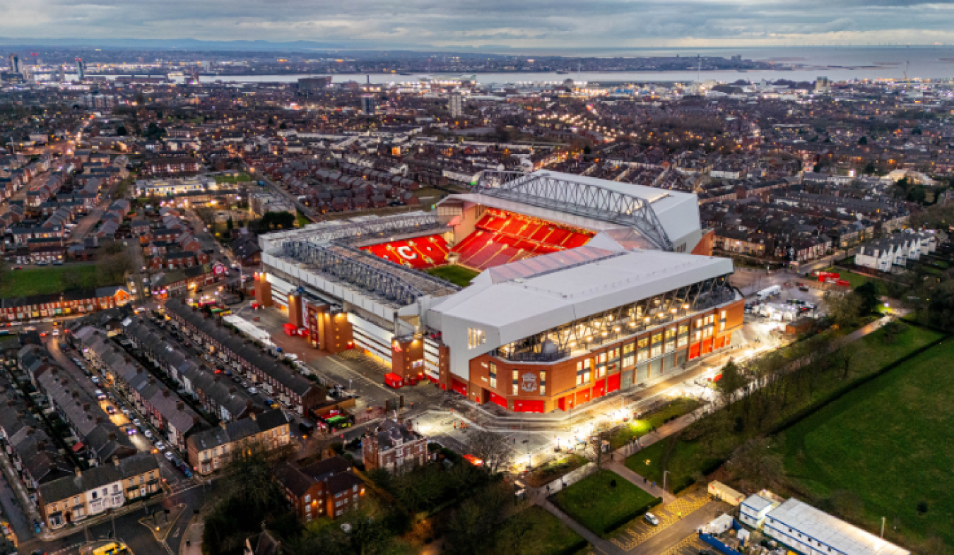 Aerial view of a large football stadium at dusk, red seating visible, surrounded by residential streets and cityscape.