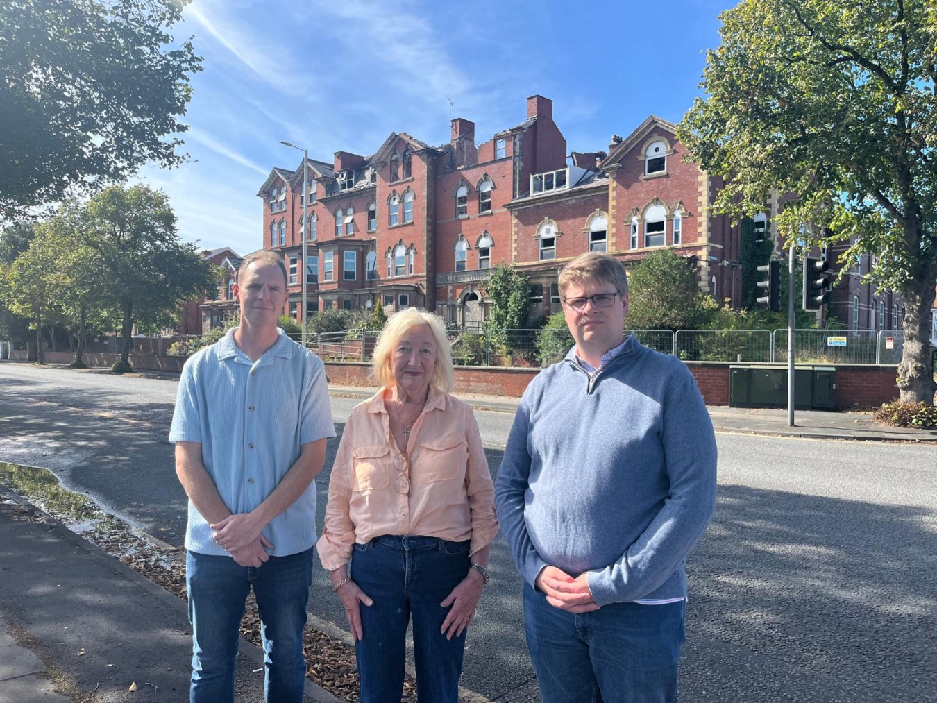 Three people stand on a street in front of a red-brick building under a blue sky.