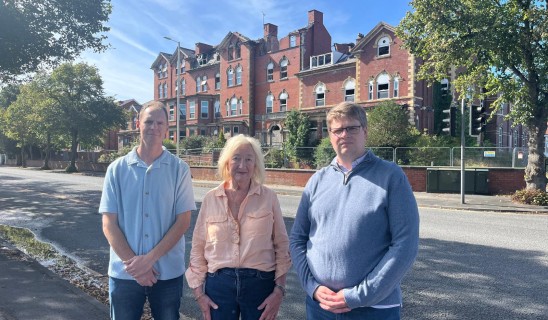 Three people stand on a street in front of a red-brick building under a blue sky.