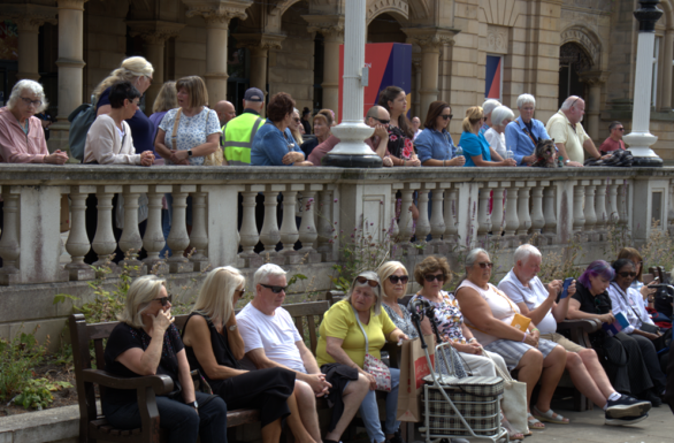 People sitting and standing along a stone balustrade courtyard, buildings in background