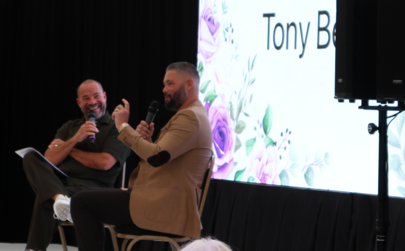 Two men seated on stage with microphones, speaker screen behind them showing floral design and partial text Tony B.