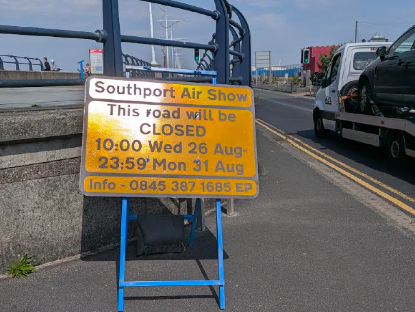 Bright yellow road sign on a blue frame near a quay, indicating closure for Southport Air Show with dates and contact info.