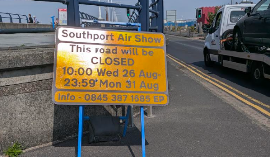 Bright yellow road sign on a blue frame near a quay, indicating closure for Southport Air Show with dates and contact info.