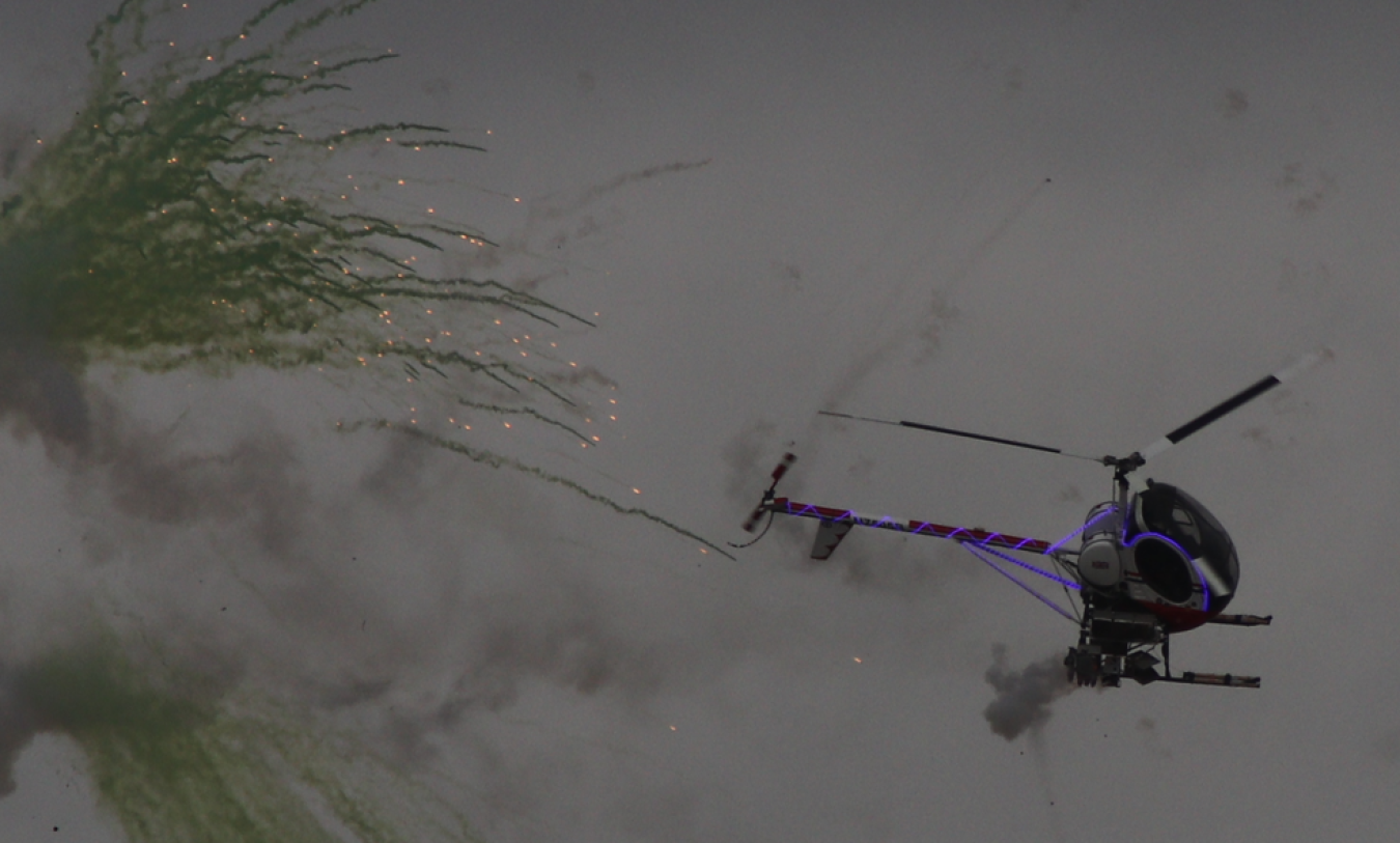 A small helicopter-like craft emits sparks and rocket-like projectiles, towing purple lines as it flies through smoky grey clouds