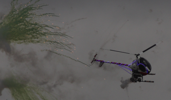 A small helicopter-like craft emits sparks and rocket-like projectiles, towing purple lines as it flies through smoky grey clouds