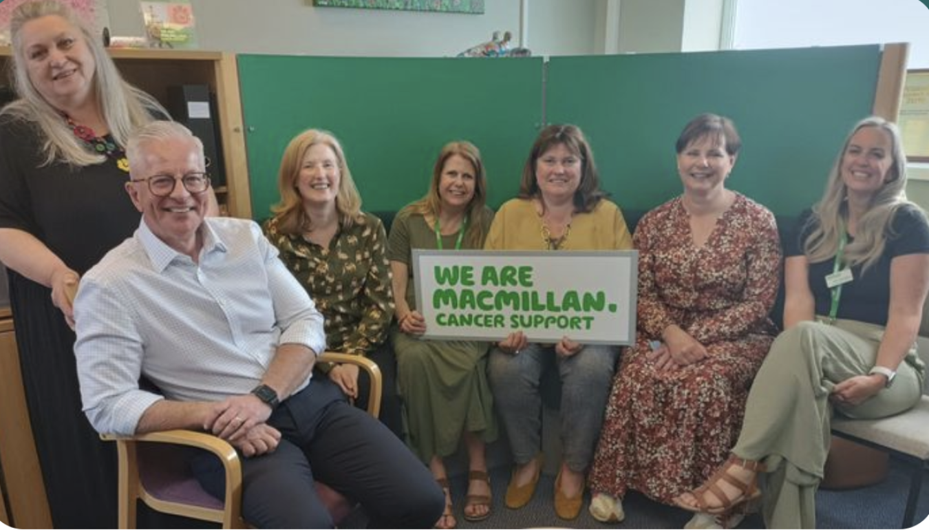 Group of six people indoors, seated and smiling, holding a sign that reads "We Are Macmillan Cancer Support".