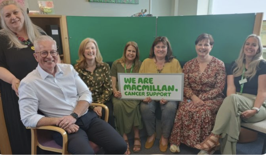 Group of six people indoors, seated and smiling, holding a sign that reads "We Are Macmillan Cancer Support".