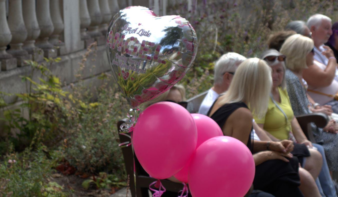 Group of people seated outdoors with pink balloons and a heart-shaped foil balloon