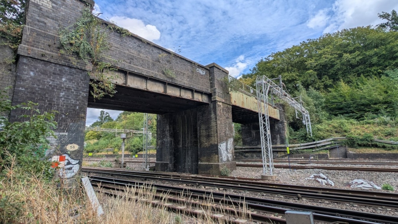 A weathered brick railway bridge over multiple tracks with ivy and graffiti, and a metal gantry to the right amid greenery