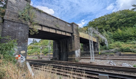 A weathered brick railway bridge over multiple tracks with ivy and graffiti, and a metal gantry to the right amid greenery