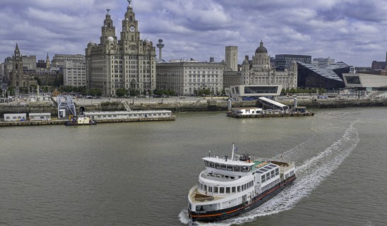 Liverpool waterfront with historic buildings along the river and a large white passenger vessel in the foreground.