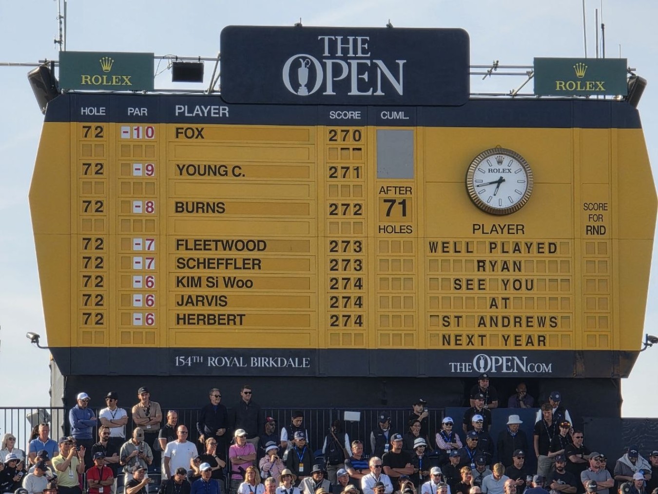 Large yellow The Open scoreboard at a golf tournament with players' names and scores, spectators below.