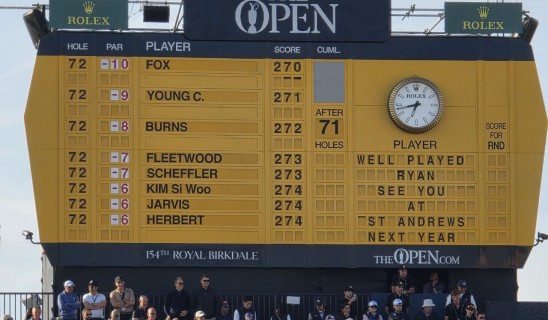 Large yellow The Open scoreboard at a golf tournament with players' names and scores, spectators below.