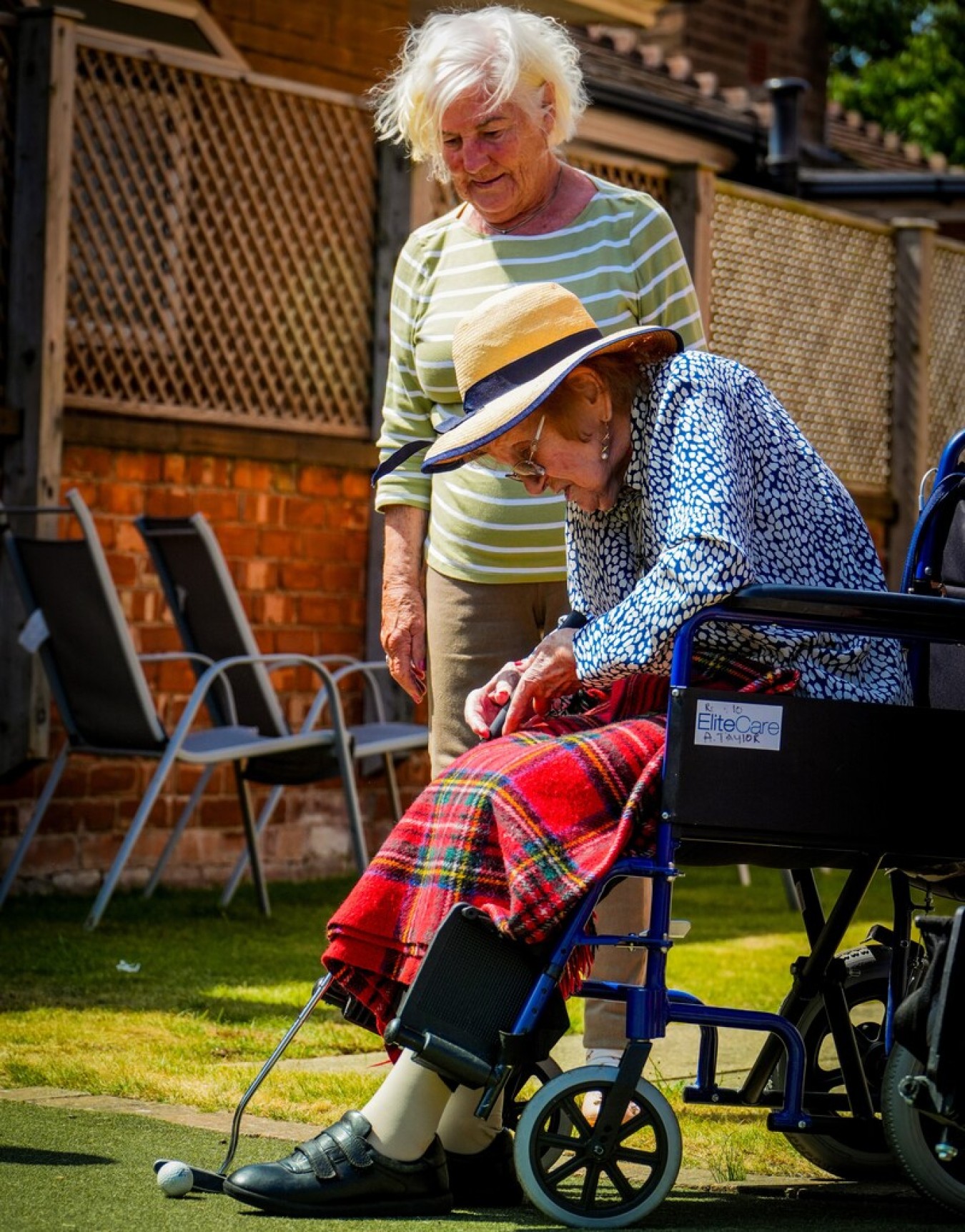 Two elderly women outdoors, one steering a wheelchair-bound companion who is about to putt a golf ball on a green.