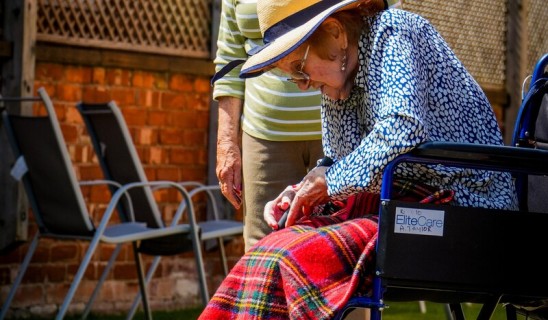 Two elderly women outdoors, one steering a wheelchair-bound companion who is about to putt a golf ball on a green.