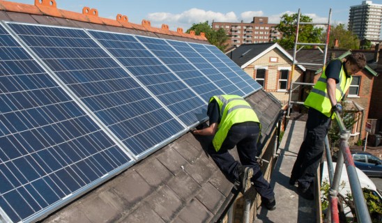 Two workers in high-visibility vests install or inspect solar panels on a sloped roof.