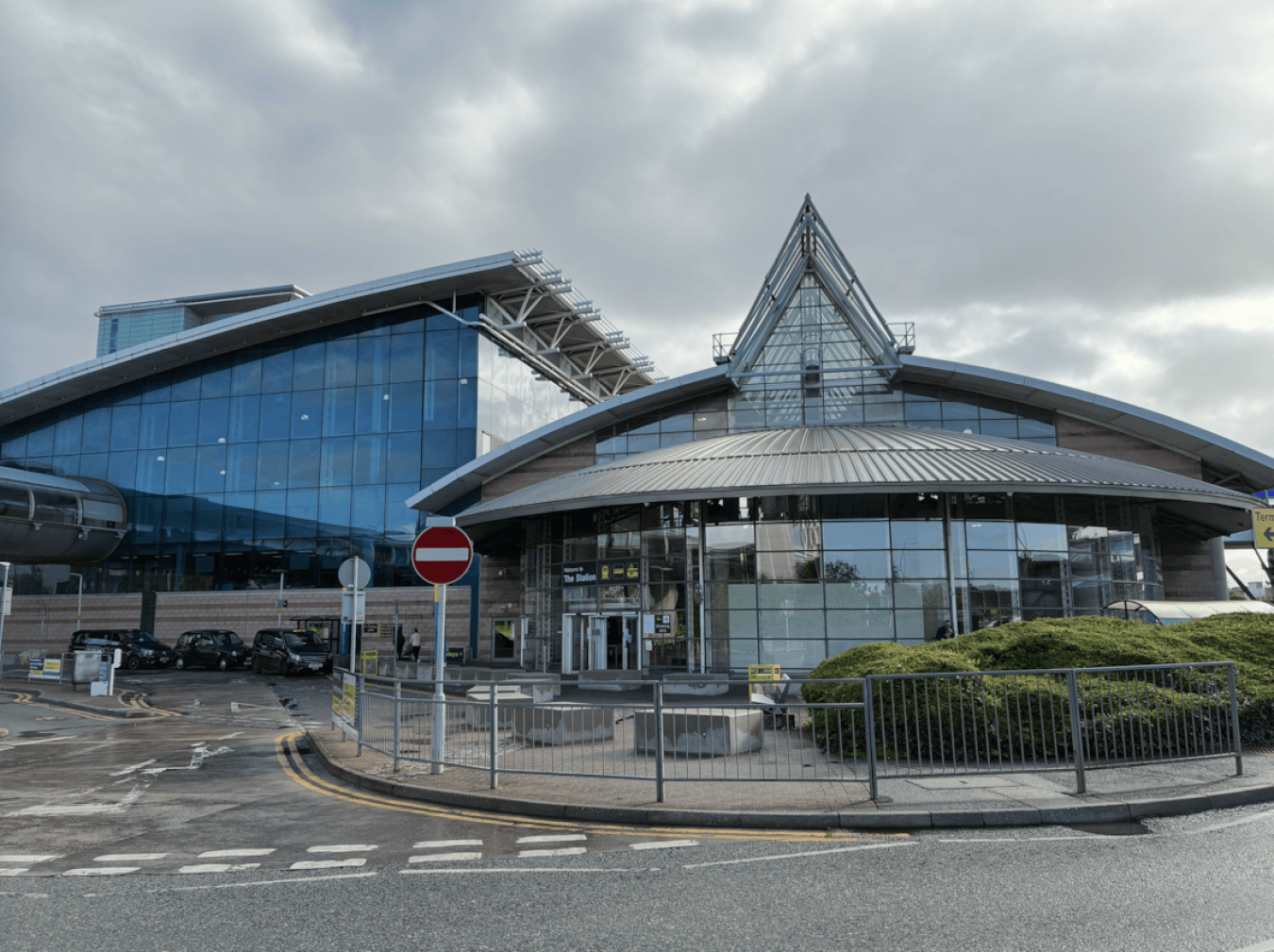 Modern glass-fronted building with curved roof and triangular glass peak, surrounded by a driveway and railings