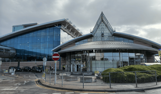 Modern glass-fronted building with curved roof and triangular glass peak, surrounded by a driveway and railings
