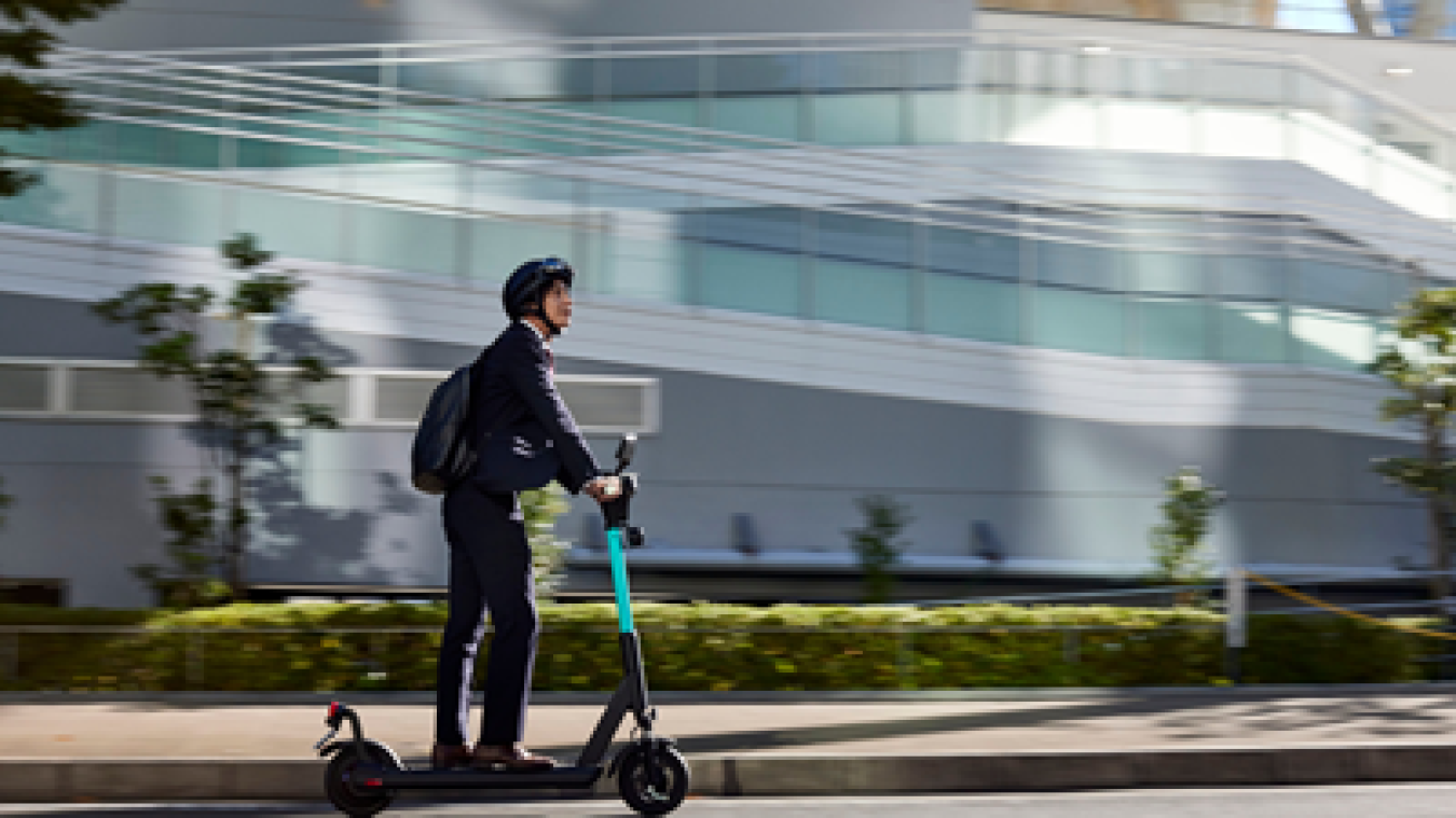 Man in business attire rides an electric scooter along city street with modern glass building backdrop