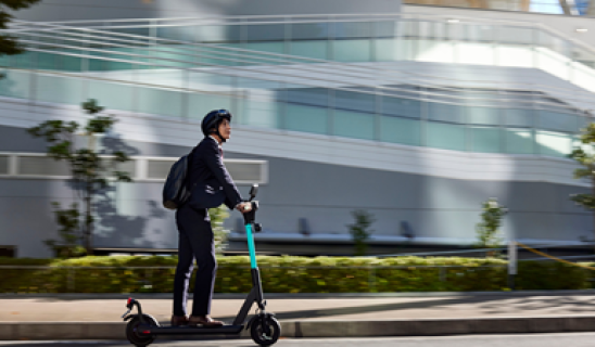 Man in business attire rides an electric scooter along city street with modern glass building backdrop