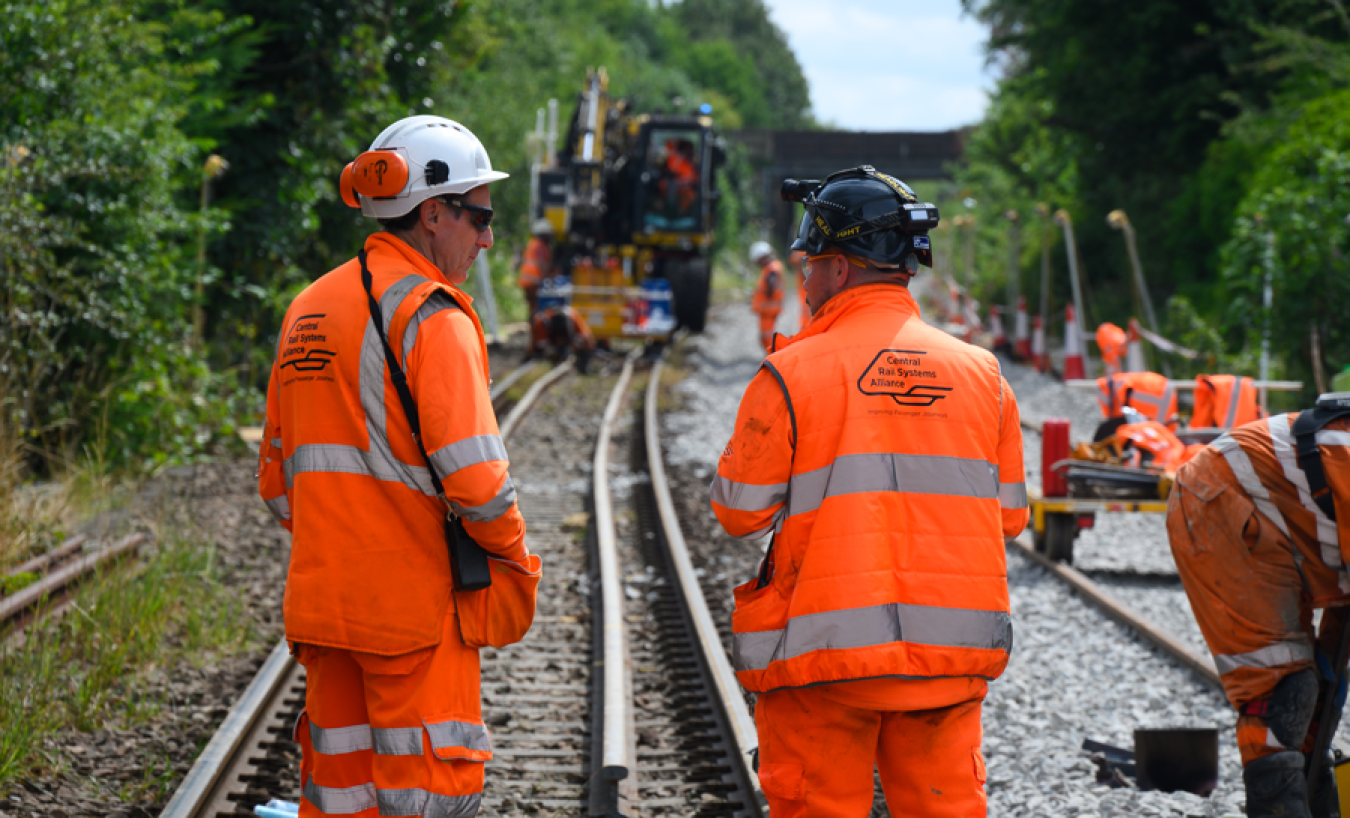 Railway workers in high-visibility orange coats on tracks, with construction equipment and greenery on the sides