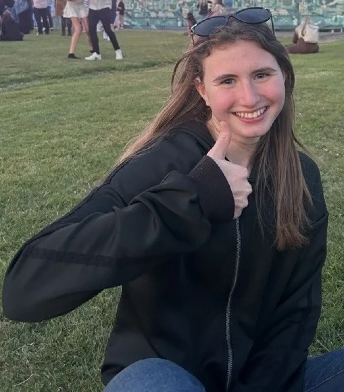 Smiling woman in dark jacket giving thumbs up while sitting on grassy field.