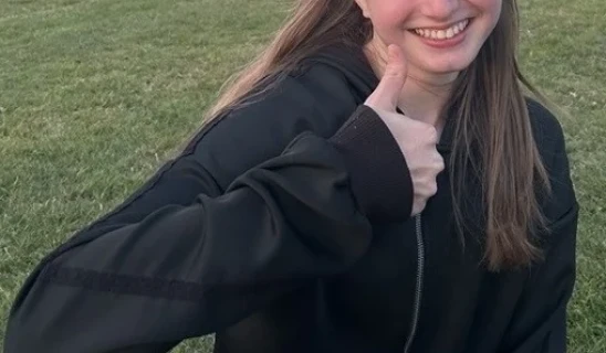 Smiling woman in dark jacket giving thumbs up while sitting on grassy field.
