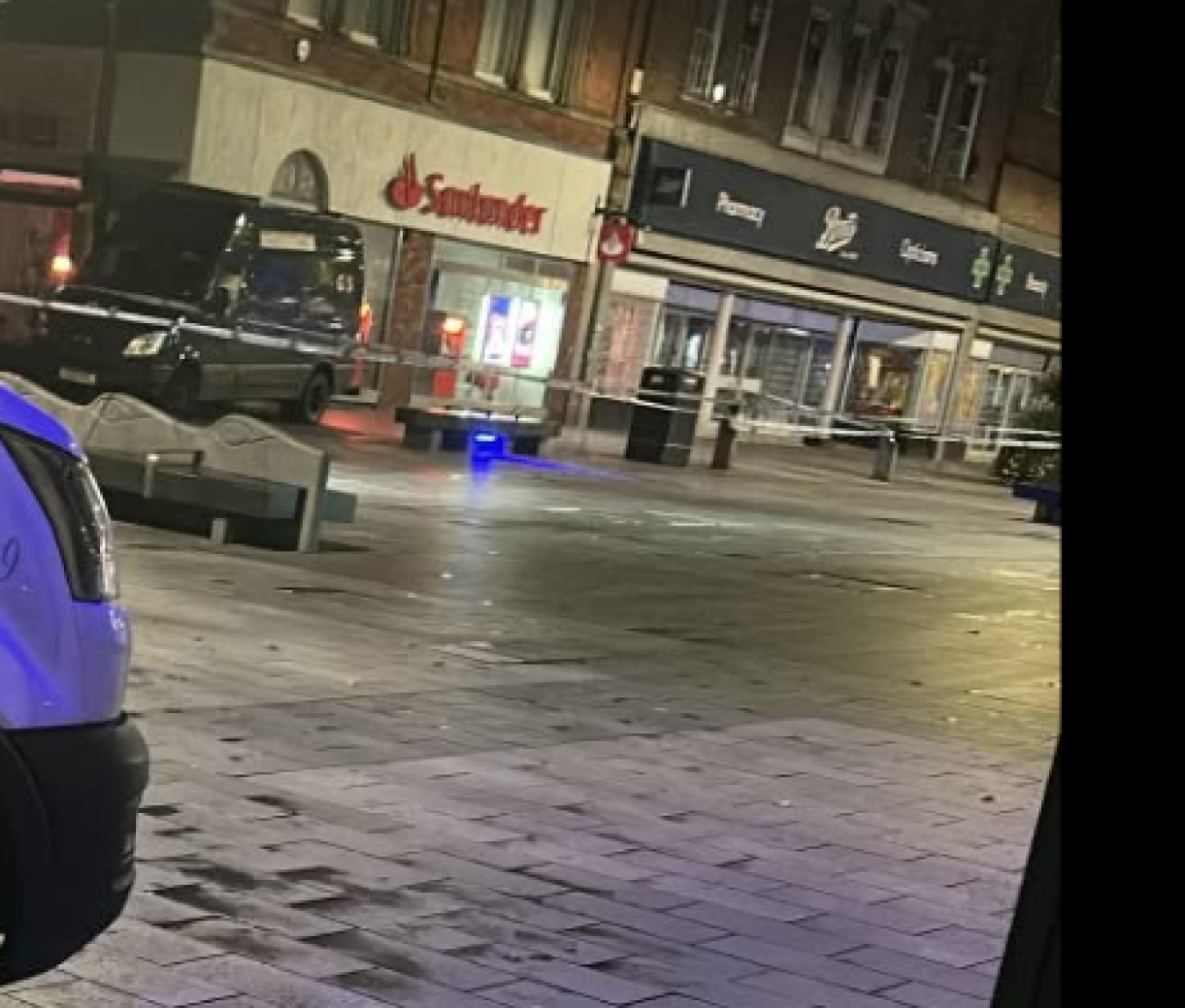 Night street scene outside shops with police tape and a few storefronts illuminated, pedestrians absent.