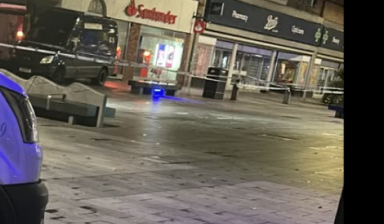 Night street scene outside shops with police tape and a few storefronts illuminated, pedestrians absent.