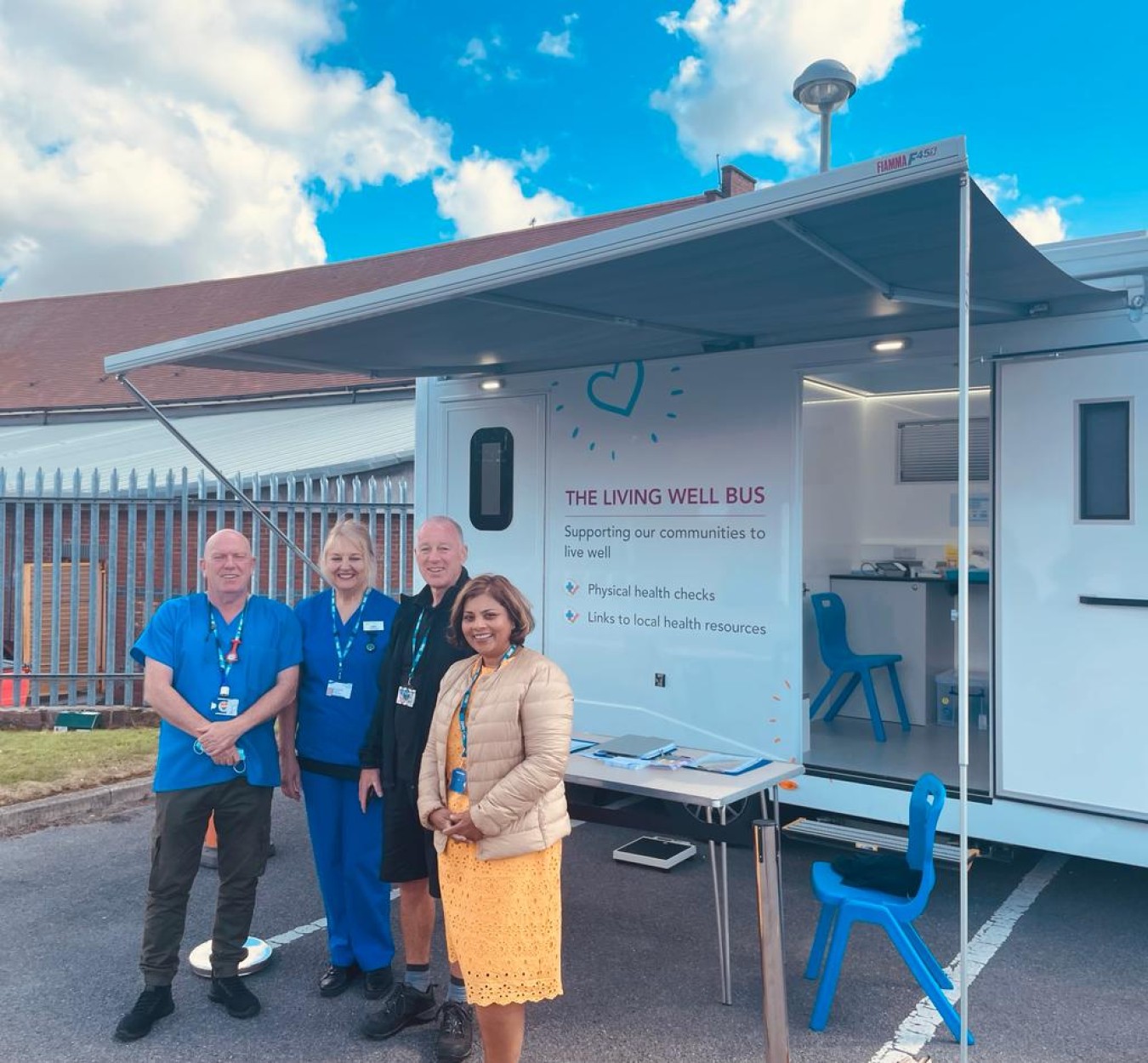 Four people stand outside a mobile health unit labelled 'The Living Well Bus' with a blue sky backdrop.