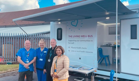 Four people stand outside a mobile health unit labelled 'The Living Well Bus' with a blue sky backdrop.