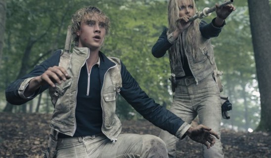 Two people in a forest setting, wearing light-colored vests and beige pants, appearing alarmed and reaching forward with props.