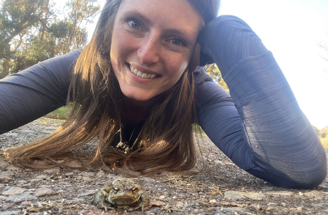 Smiling woman lying on the ground next to a small frog on a rocky path.