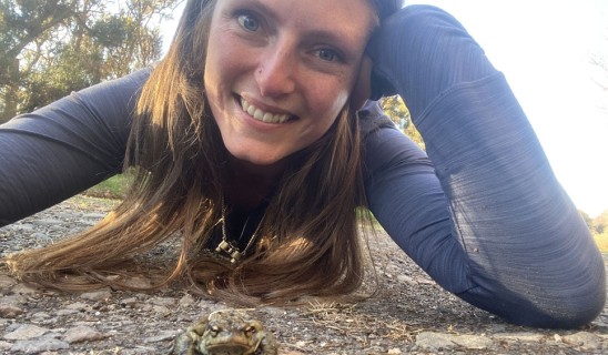 Smiling woman lying on the ground next to a small frog on a rocky path.